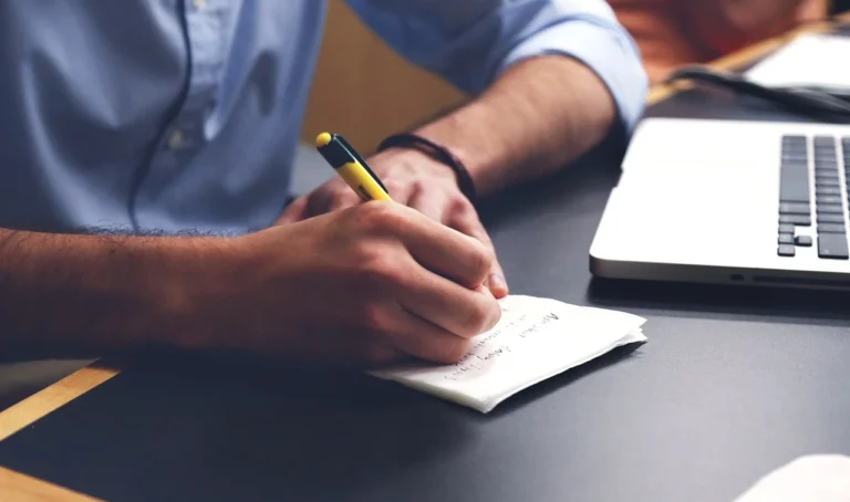 Pessoa escrevendo em um caderno com laptop ao lado, ambiente de trabalho organizado e moderno.